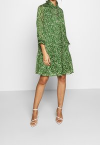 Abito lungo verde, fluido, fino al ginocchio con un motivo floreale e maniche lunghe. Presenta una chiusura frontale con bottoni e una vestibilità comoda. Indossato con tacchi bianchi a griglia.