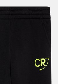 Čierne teplákové nohavice vyrobené z mäkkej látky, s neonovo žltým logom "CR7" a akcentom v podobe Swoosh od Nike. Elastický pás so prešitými detailmi.