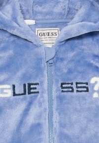 Svijetloplavi flis zip-up dukserica s kapuljačom s natpisom "GUESS?" u tamnoplavoj i bijeloj boji, mekane teksture. Brand oznaka vidljiva unutra.