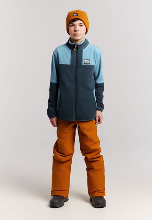 Jonge persoon draagt een blauwe en donker turquoise fleecetrui, een oranje muts, oranje skibroek en zwarte laarzen, staand en naar voren gericht.