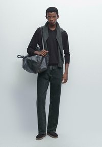 Schwarzer Pullover, darüber ein grauer Schal, dunkle Jeans und braune Slipper. Schwarze Ledertasche in der linken Hand gehalten.