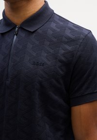 Homme portant un polo bleu foncé texturé avec une fermeture éclair et un logo brodé "BOSS" sur la poitrine.