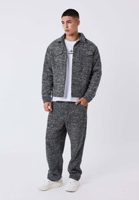 Giovane uomo che indossa una giacca a maglia grigia e pantaloni abbinati su una t-shirt bianca con grafica, abbinato a sneakers bianche, in piedi contro uno sfondo semplice.