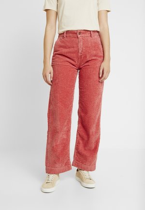 Personne portant un pantalon en velours côtelé rose taille haute à jambes larges, des baskets beiges et une chemise beige à manches courtes sur un fond uni.