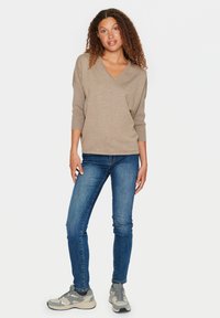 Bež pulover z V-izrezom in 3/4 rokavi, oblečen s sploščenimi modrimi jeans hlačami in sivimi športnimi čevlji. Gladka tekstura z rebrastimi detajli na rokavih.