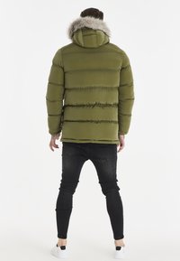 Olivengrøn pufferjakke med hætte og faux fur trim, ribbet tekstur og vandret syninger. Båret med sorte stramme jeans.