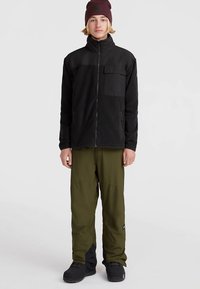 Noor inimene, kes kannab musta lukujope, oliivrohelisi pükse, musti kingi ja tumedat burgundia mütsi, seisab ühevärvilise heleda tausta ees.