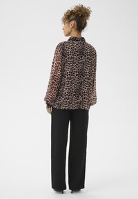 Moteris su garbanotais plaukais sukasi į kuodą, vilkinti leopardų rašto ilgas rankoves turintį bliuską ir juodas plačiašlaistes kelnes, stovi atsisukusi nuo paprastos fono.