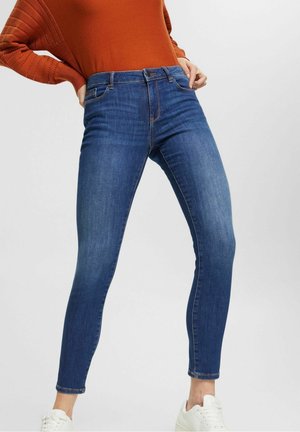 Osoba ubrana w obcisłe niebieskie dżinsy, rdzawy długi rękaw i białe trampki, stojąca z rękami na biodrach na jednolitym tle.