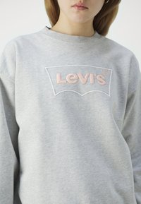 Jeune personne portant un sweat-shirt gris clair avec le logo Levi's brodé en rose pâle sur la poitrine, visible des épaules jusqu'au milieu du torse.