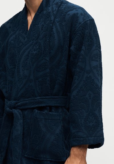 Personne portant un peignoir texturé bleu foncé avec une ceinture nouée à la taille et une poche avant visible sur le côté droit.