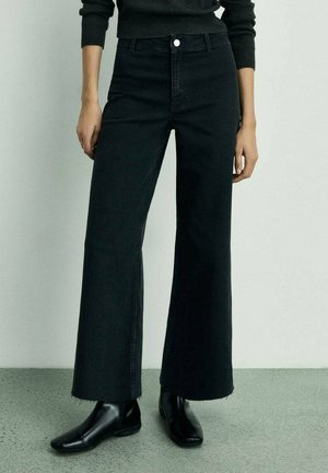 Black wide-leg jeans in denim with a high waist, featuring a front button closure, clean lines, and raw hem details. Paired with black boots.