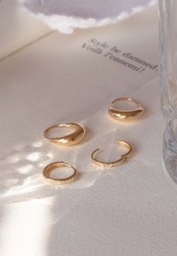 Vier goldene Ringe auf strukturiertem Papier: ein polierter Dom, zwei schlichte Bänder und ein facettierter Ring. Ein Glas und ein Stoff sind ebenfalls vorhanden.