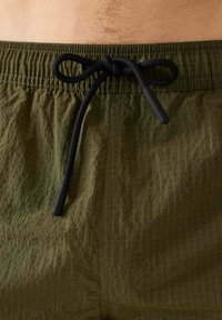 Olijfgroene zwembroek met een gestructureerde stof, een elastische tailleband en een zwarte trekkoord met accent uiteinden. Ruitpatroon zichtbaar.