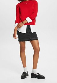 Pull rouge texturé porté par-dessus une chemise blanche, associé à une jupe en jean noire avec un ourlet effiloché et des chaussures noires à semelle épaisse.