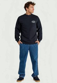 Zwarte sweatshirt met een ronde hals en een klein grafisch detail op de borst, gecombineerd met blauwe spijkerbroek en zwarte sneakers.