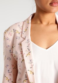 Blazer med blomsterprint i lyserødt stof med tekstureret overflade. Har et spidst krave og bæres over en hvid V-udskæring top.