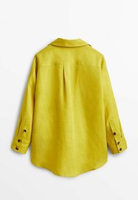 Keltainen pellava-paita, jossa on kaulus, pitkät hihat ja napilla kiinnitettävät hihansuut. Sisältää yhden takataskun ja suoran helman.