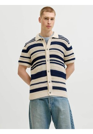 Jeune homme portant une chemise en maille à manches courtes rayée crème et marine, boutonnée, par-dessus un t-shirt bleu clair et un jean bleu clair.