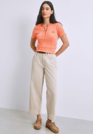 Mujer de pie con las manos detrás de la espalda, vistiendo un top de punto coral de manga corta, pantalones anchos beige y zapatos marrones con cordones.