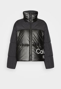 Svart puffjacka med hög krage, glänsande frampanel, matta ärmar och stor vit logotext. Dragkedjestängning framtill i mitten.