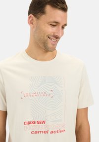 Beige bomulds t-shirt med et grafik med blå linjer og rød tekst. Teksten inkluderer "UNLIMITED ADVENTURER" og "CHASE NEW HORIZONS."