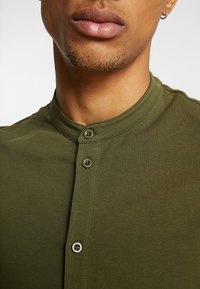 Olijfgroene, korteg mouwen shirt met een ronde halslijn, voorzien van een knoopsluiting met twee knopen van donker kunststof. Gladde textuur.