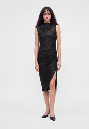 Le modèle se tient devant un fond de studio uni, vêtue d’une robe midi noire sans manches à la texture travaillée, fendue sur le côté, et de chaussures noires à talons.