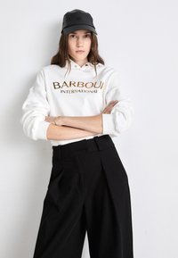 White hoodie with gold "BARBOUR INTERNATIONAL" print, paired with high-waisted black trousers. Model wears a black cap. Backdrop is neutral.