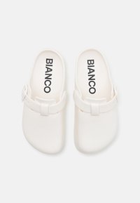 Hvide slip-on klipklapper lavet af blødt materiale, med en strop med en knapdetalje og mærkenavnet "BIANCO" trykt indeni.