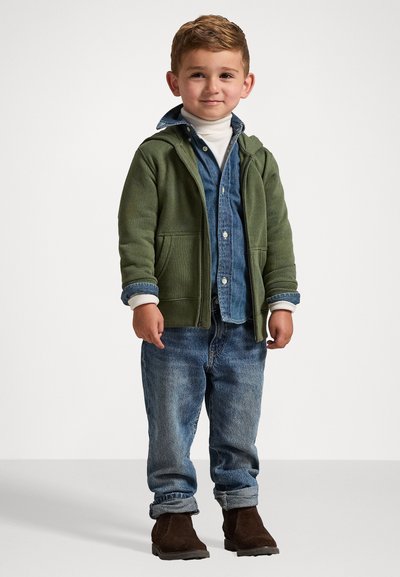 Zelená mikina na zip, modrá denimová košile na knoflíky, krémový rolák, modré džínsy, hnědé kotníčkové boty; neformální vrstvený outfit pro dítě.