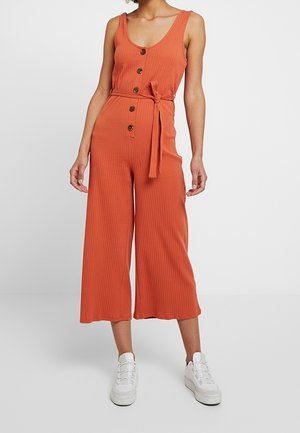 Woman wearing a sleeveless burnt orange buttoned jumpsuit with a waist tie and wide-leg cropped pants, paired with white sneakers.