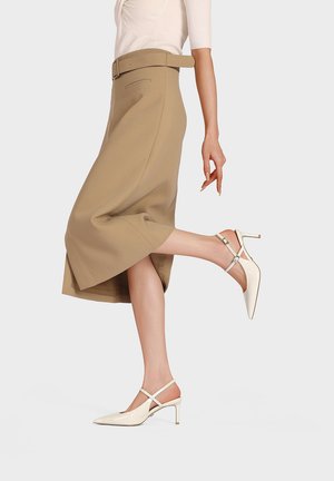 Mujer con falda midi beige con cinturón, camiseta blanca de manga corta y zapatos de tacón de charol crema con tira trasera, posando con una pierna levantada.