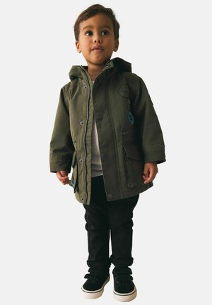 Noor laps, kes kannab oliivirohelist kapuutsiga mantlit laigukestega, musti pükse ja musti tossusid, seisab ja vaatab veidi kõrvale.