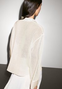 Cardigan beige translucide avec une coupe décontractée, col et manches longues. Texture en maille fine, ourlet arrondi, et superposé sur un vêtement léger.