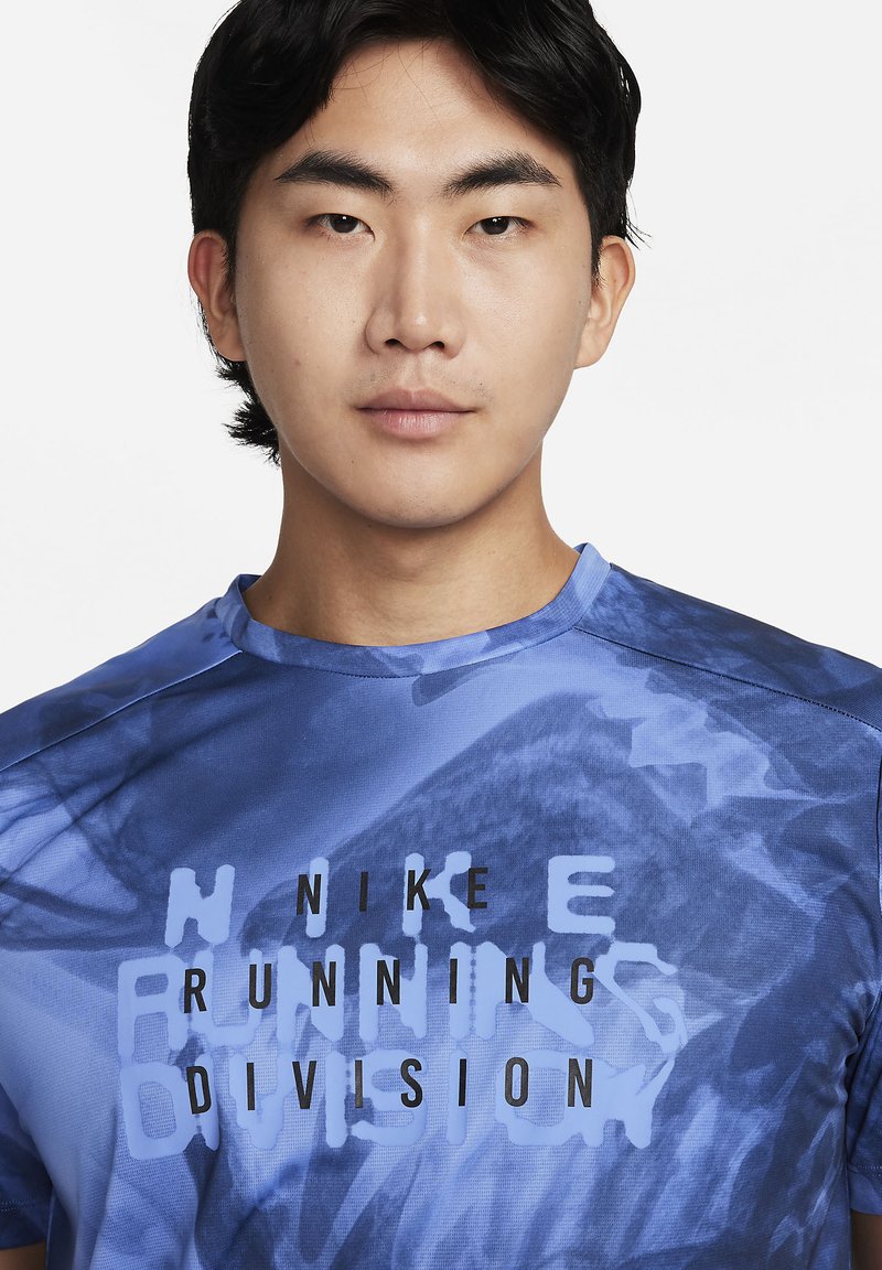 Jeune homme avec des cheveux noirs courts portant un t-shirt bleu de la division course Nike avec un motif tie-dye sur fond blanc.