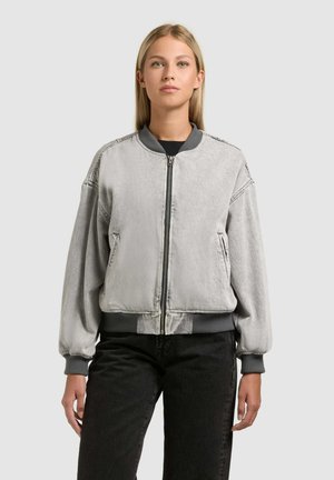 Jonge vrouw draagt een lichtgrijs bomberjack met ritssluiting en geribbelde boorden en kraag, gecombineerd met een zwarte broek, staand tegen een effen achtergrond.