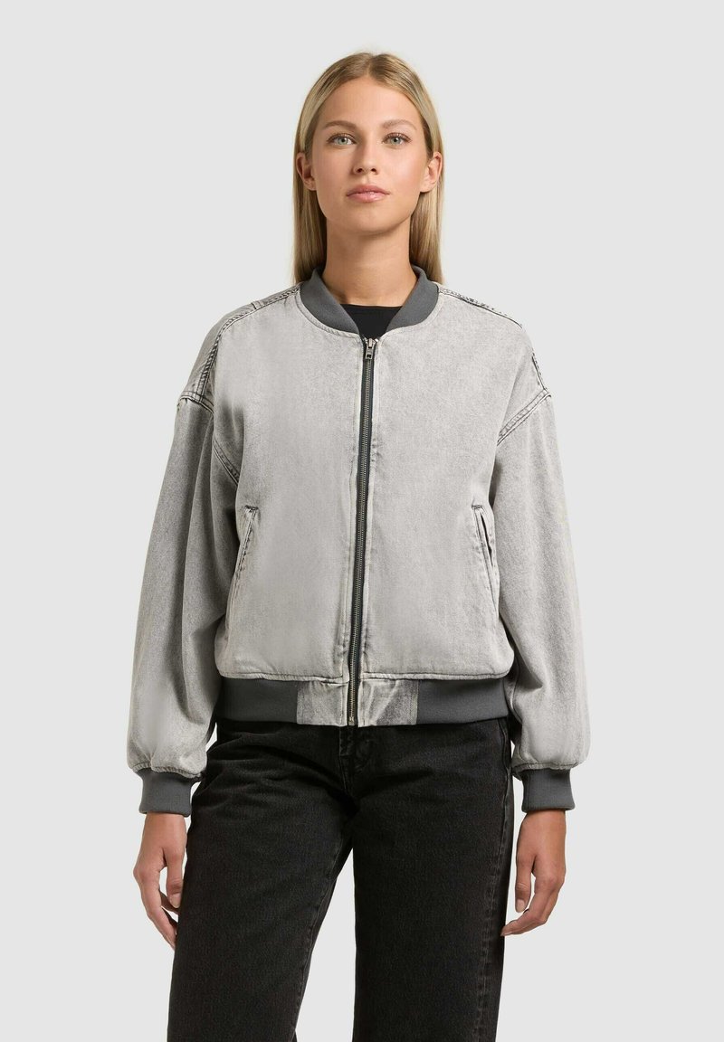 Jeune femme portant une veste bomber grise claire à fermeture éclair, avec des poignets et un col côtelés, associée à un pantalon noir, debout devant un fond uni.