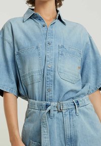 Ljusblå denimskjorta med korta ärmar, två bröstfickor, knäppning fram och ett matchande bälte. Mjuk textur och avslappnad passform.