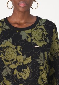 Sweat-shirt noir avec un motif floral vert, col rond et détails en métal doré sur la poitrine. Texture de tissu douce.