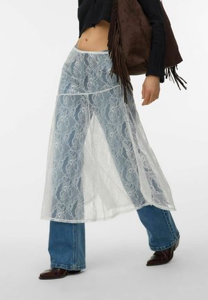 Personne portant un jean évasé bleu sous une longue jupe blanche en dentelle transparente, des chaussures sombres à bouts pointus, un haut noir court et tenant un sac à franges marron.