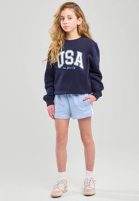 Felpa marina con grafica "USA New York City", abbinata a pantaloni corti azzurri chiari e calze bianche. Le sneaker hanno accenti beige.