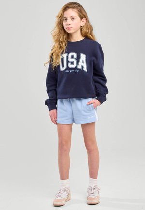 Navy collegepaita, jossa on "USA New York City" -grafiikka, yhdistettynä vaaleansiinisiin shortseihin ja valkoisiin sukkiin. Kengissä on beigejä yksityiskohtia.