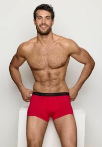 Boxer ajustados de algodón rojo con cinturilla negra que presenta el logotipo de la marca, de diseño ceñido y textura suave. El modelo posa con un fondo neutro.