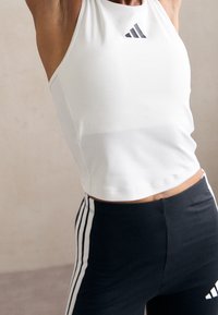 Gros plan d'une personne portant un haut sans manches blanc Adidas et un pantalon noir avec des rayures blanches, posant les bras levés.