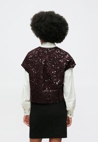 Ženska s afro frizuro nosi belo bluzo z dolgimi rokavi in kratko črno krilo z temno vijoličastim bleščečim kratkim jopičem, gledano od zadaj.