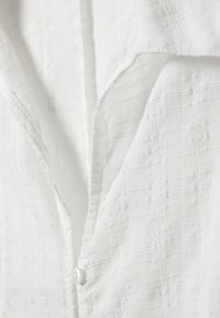 Camicia bianca con tessuto testurizzato caratterizzato da sottili linee verticali, colletto classico e chiusura con un solo bottone sul petto.