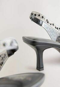 Schwarze High-Heel-Sandalen mit glänzender Oberflächenbeschichtung, ausgestattet mit einem klaren Riemen, der mit schwarzen und silbernen Nieten verziert ist, sowie einer strukturierten Innensohle.