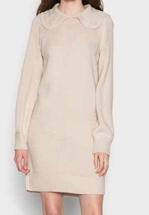 Frau trägt ein cremefarbenes Strickkleid mit rundem Kragen und langen Ärmeln vor einem schlichten hellen Hintergrund.