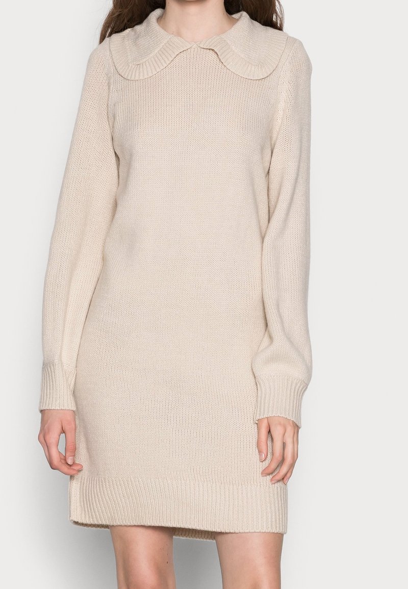 Femme portant une robe-pull en maille crème avec un col arrondi et des manches longues sur un fond clair uni.